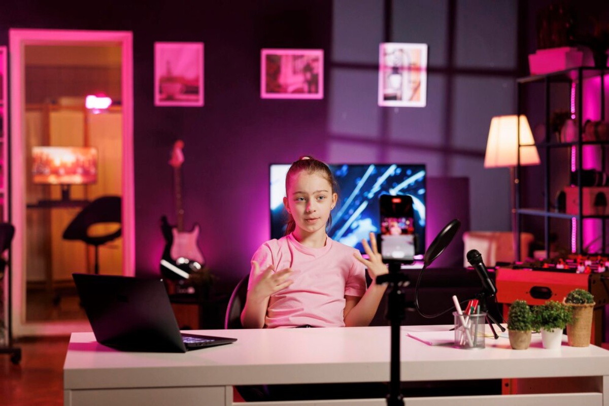 A young person in a pink shirt gestures while seated at a desk with a laptop, microphone, and potted plants in a colorful, modern room.