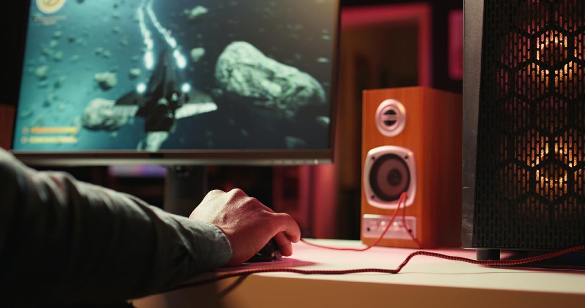 A hand controls a gaming mouse beside speakers, with a vibrant monitor displaying a spaceship navigating an asteroid field.