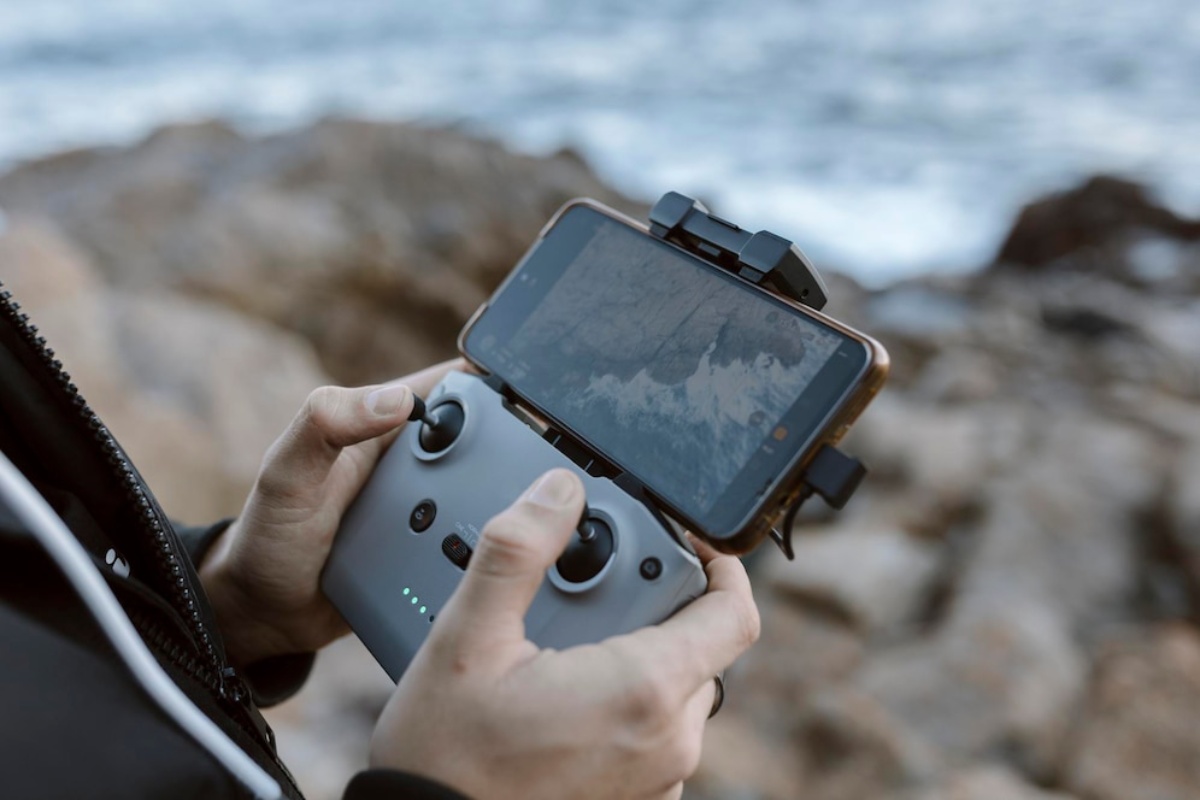 A person holds a drone controller with a smartphone mounted, displaying a landscape, against a backdrop of rocky shoreline and ocean.