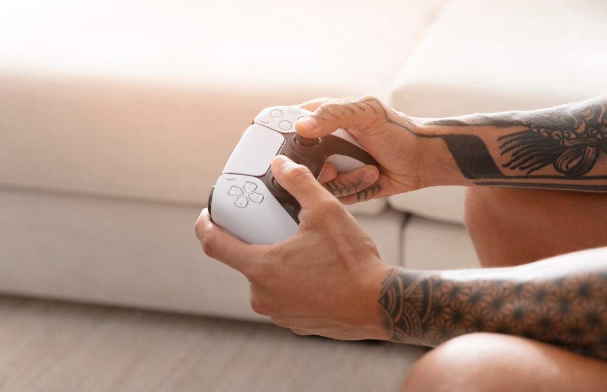 A person with tattooed arms holds a PlayStation 5 controller, focused on gaming in a cozy living room setting.