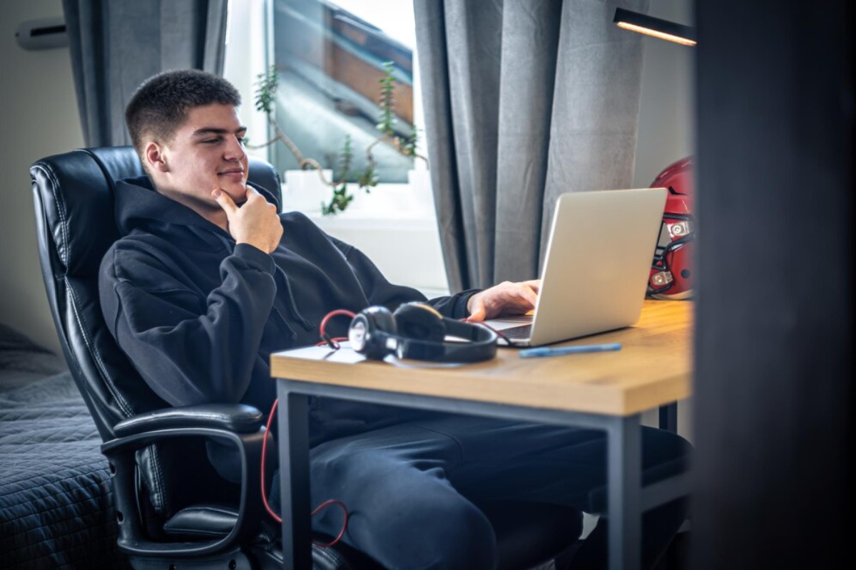 A young individual in a black hoodie sits in a chair, focused on a laptop, with headphones resting on a wooden desk.