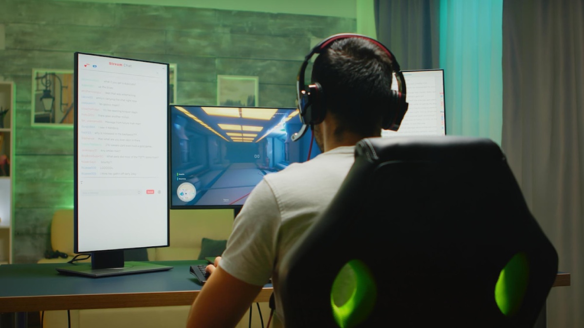 A gamer in a headset sits in front of dual monitors, one displaying gameplay and the other showing live chat messages.