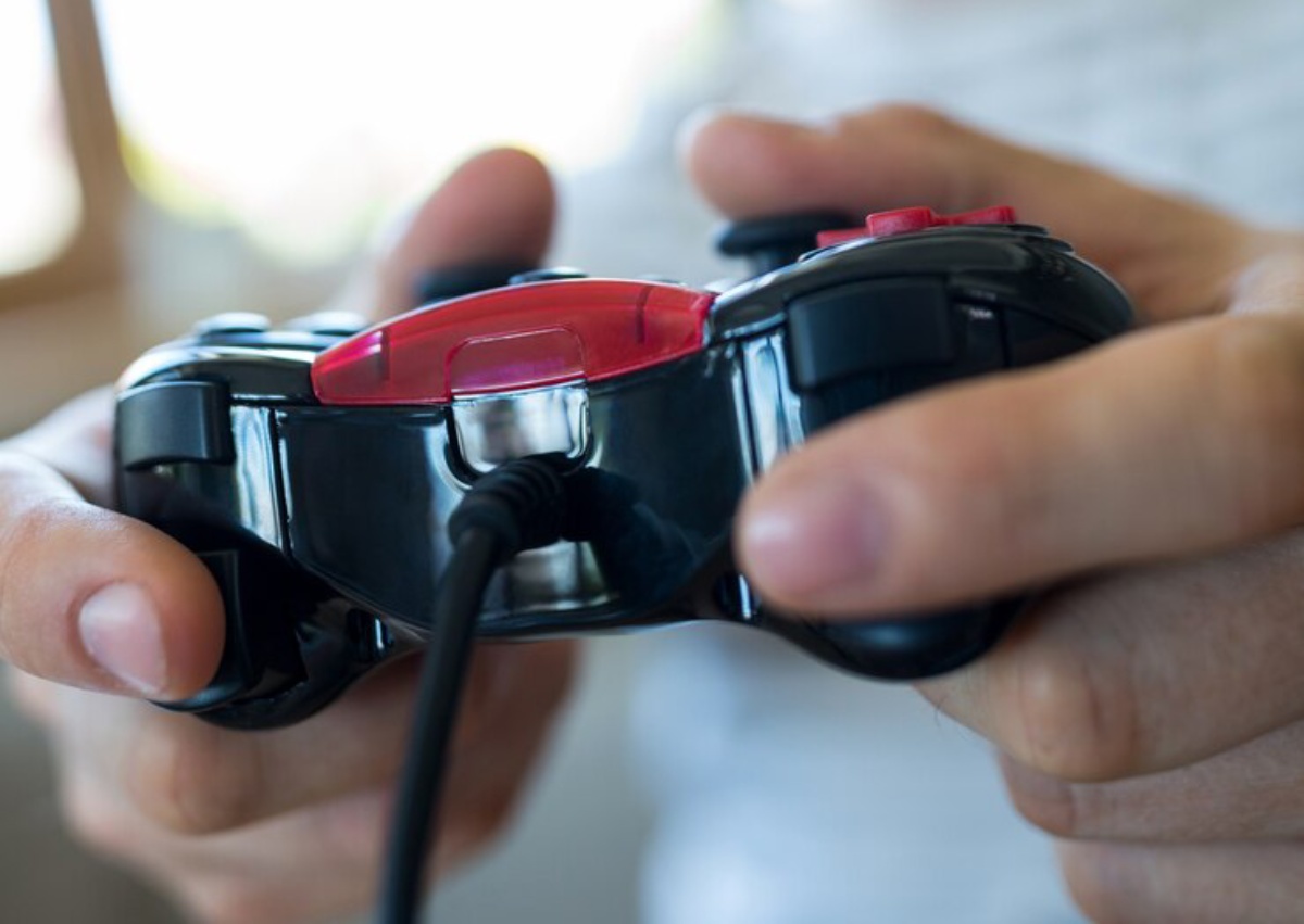 A person holds a black and red wired game controller, focused on the device with fingers poised to play.