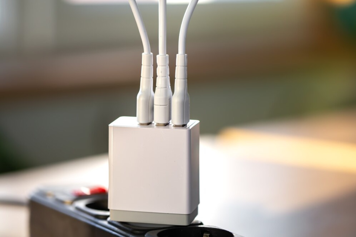 A white multi-port charger with three connected cables sits on a power strip, illuminated by natural light from a nearby window.