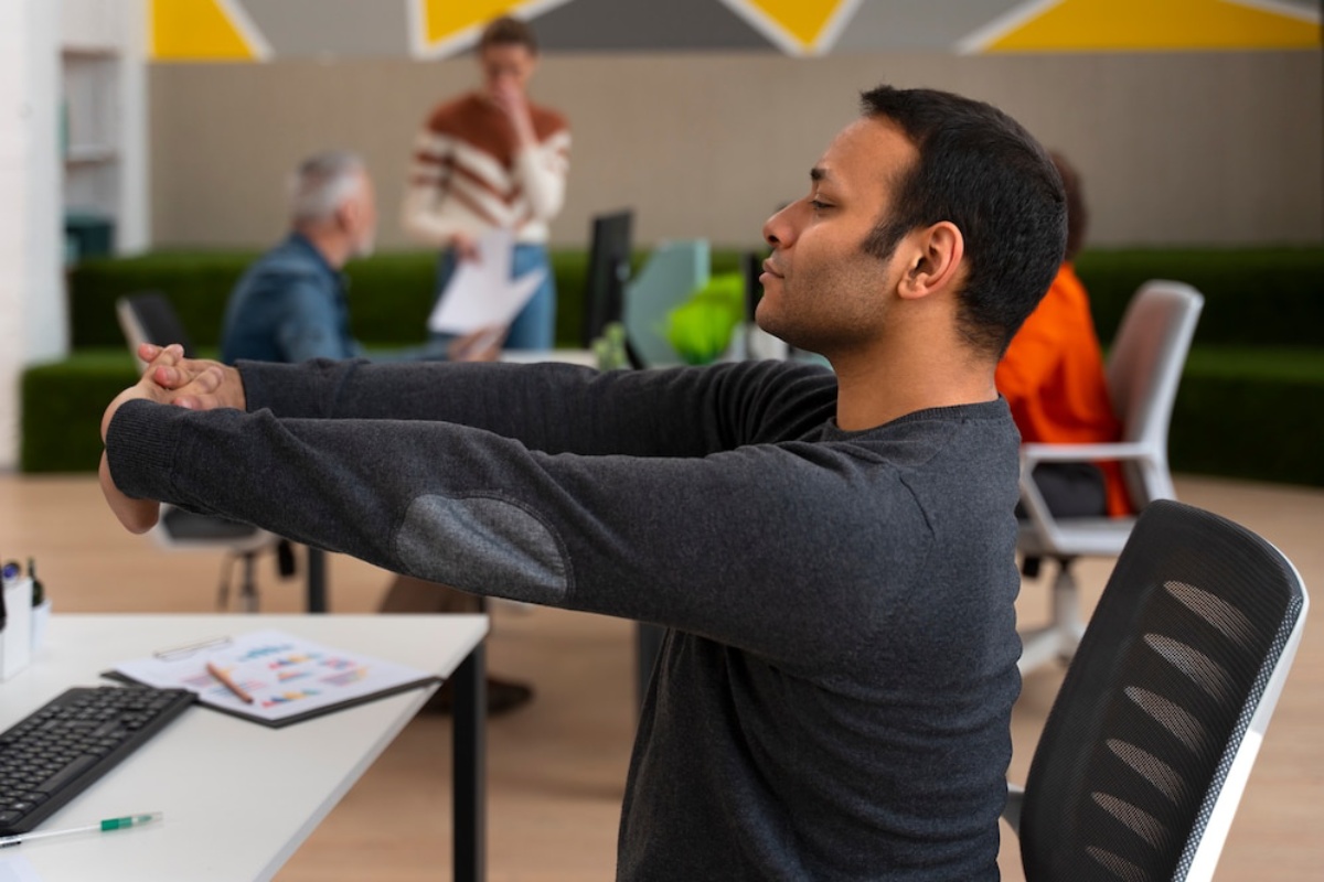 A man in a gray sweater performs a seated arm stretch at a modern office, with coworkers engaging in discussions in the background.