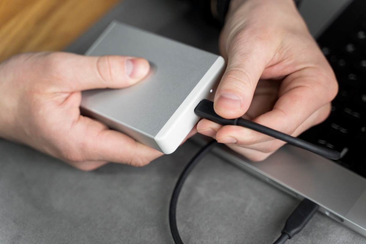 A person connects a black USB cable to a sleek silver external hard drive beside a laptop on a grey surface.