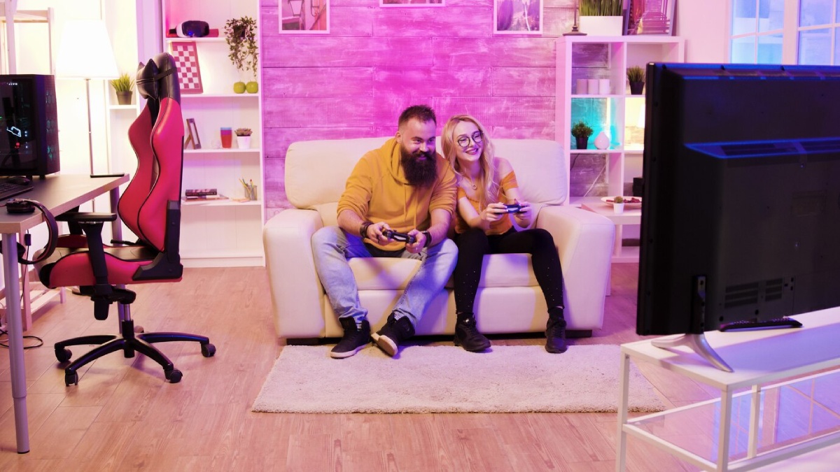 A cozy gaming setup features two people sitting on a white couch, focused on video games, with a colorful, modern room in the background.
