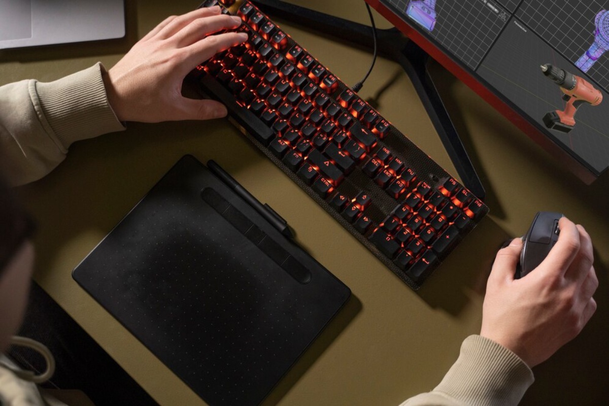 A person uses a gaming keyboard with red backlighting, a mouse, and a drawing tablet, while working on a computer.