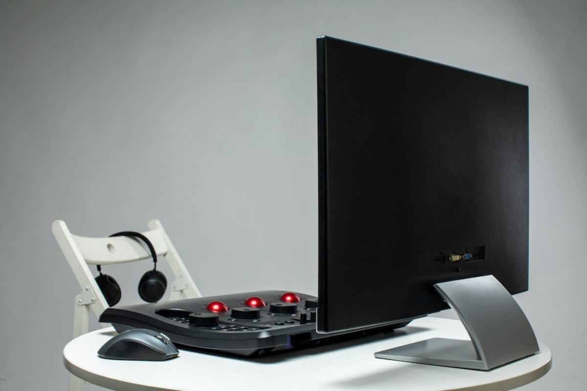 A sleek black computer monitor on a white table with a mouse, colorful buttons, and headphones on a chair beside it.