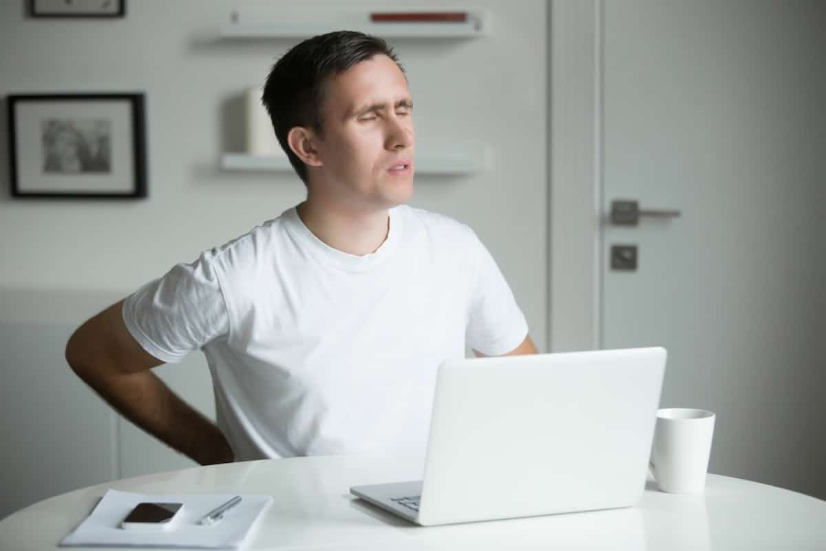 A person sits at a table with a laptop, a cup, and a plate with a small dessert, in a modern, minimalistic room.