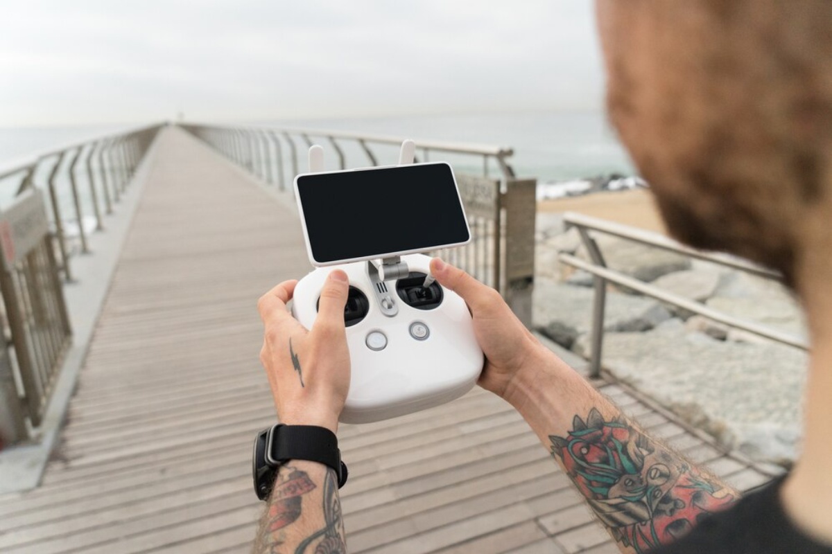 A person holds a white drone controller with a phone mounted on it, standing on a wooden pier with a cloudy ocean background.