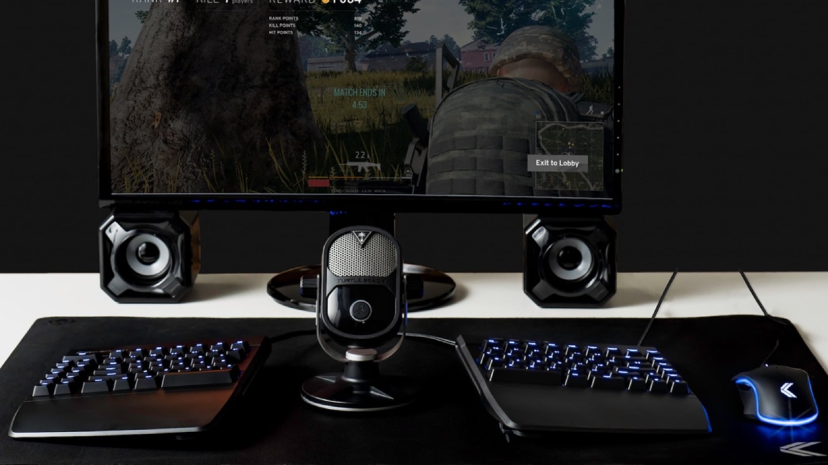 A gaming setup featuring a monitor displaying a player in action, a Turtle Beach microphone, a keyboard, and speakers.