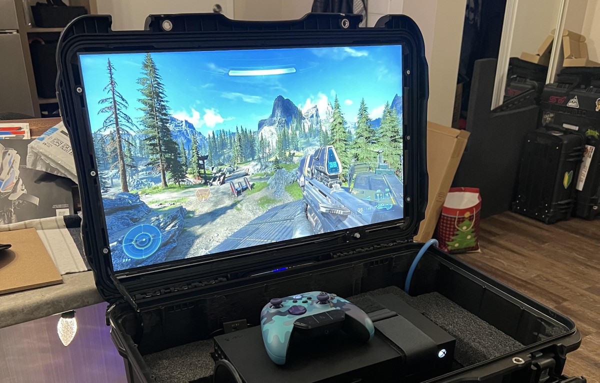 A portable gaming setup featuring an Xbox with a controller and a monitor displaying gameplay from Halo Infinite amidst a scenic landscape.